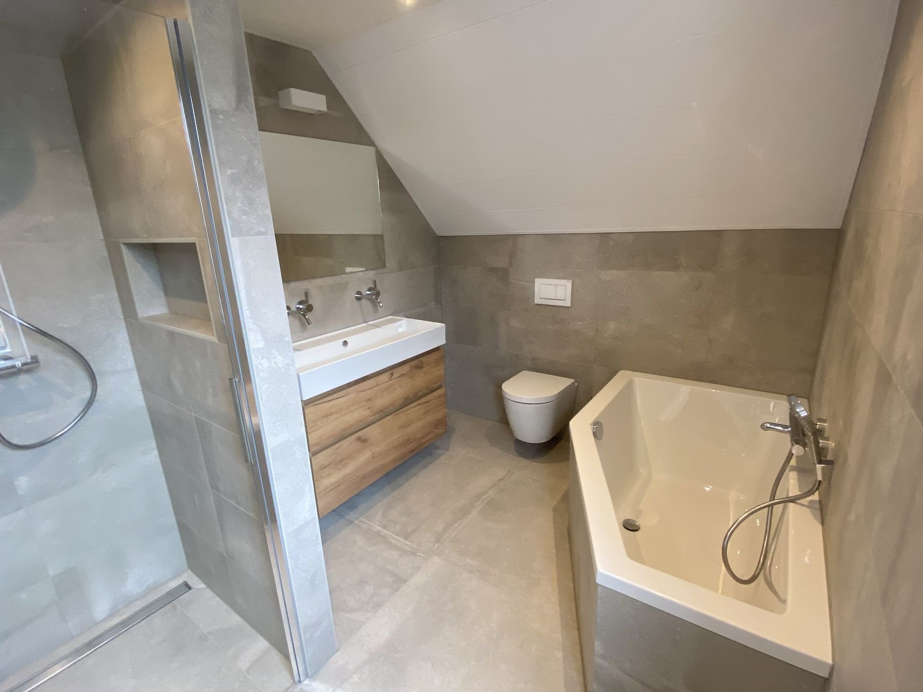 Badkamer met grote grijze tegels, douche met nis, dubbel badmeubel en een hoekbad onder een schuin dak