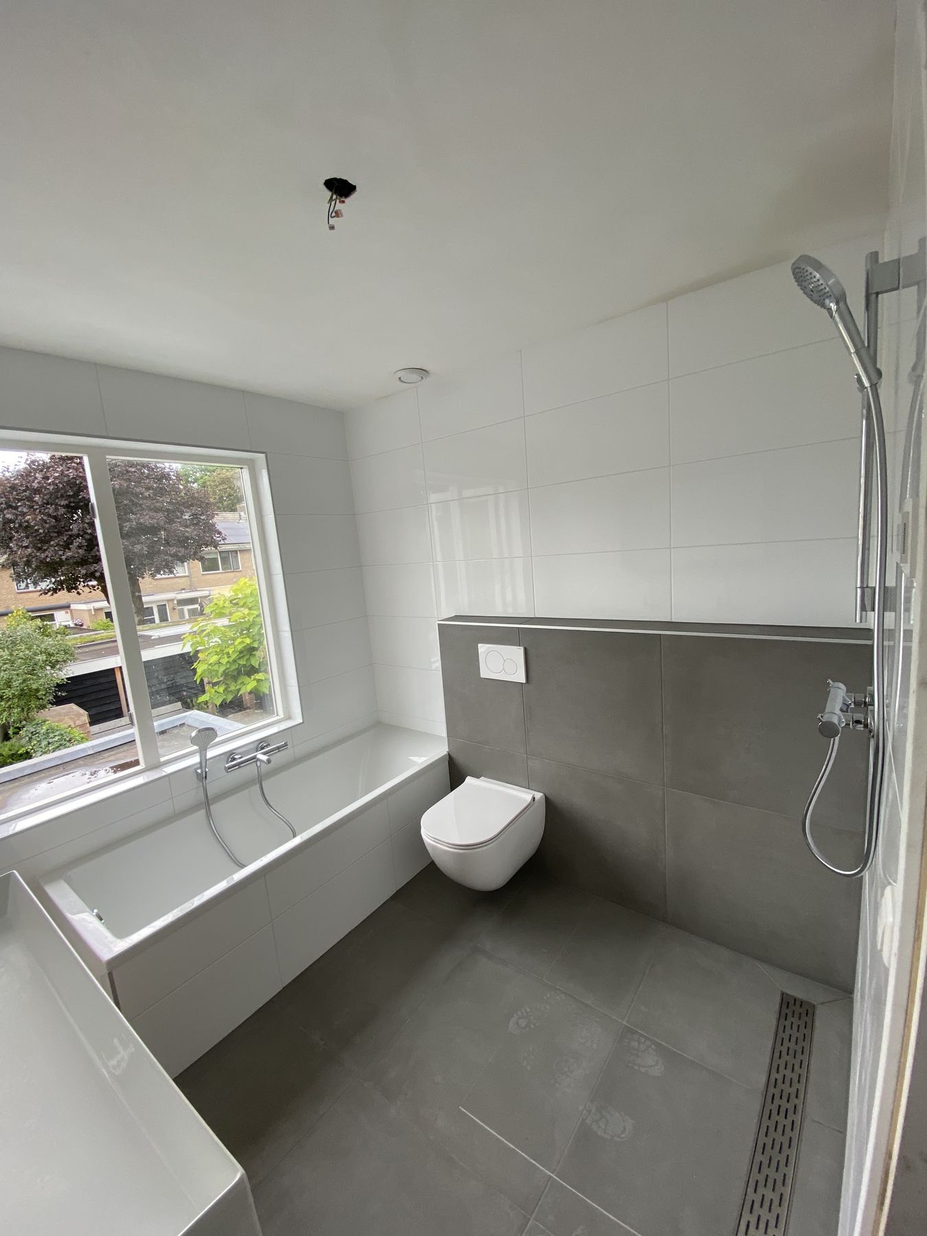 Badkamer met witte wandtegels, donkere vloer, ingebouwd bad onder het raam en een wandcloset