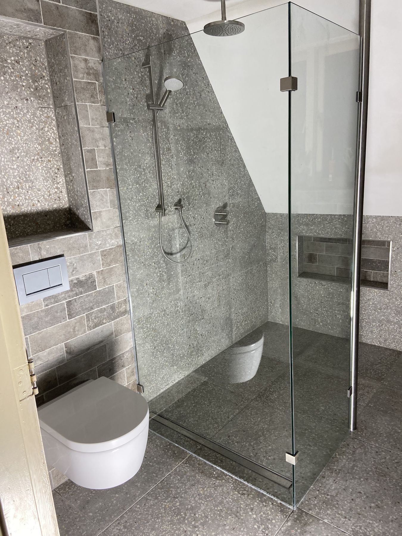 Badkamer met grijze terrazzotegels, glazen douchewand, nis en wandcloset onder een schuin dak
